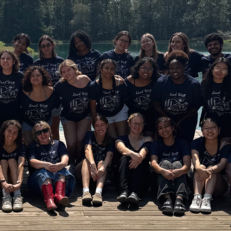 Photograph of a large group of smiling young adults in matching shirts posing outdoors by a lake.
