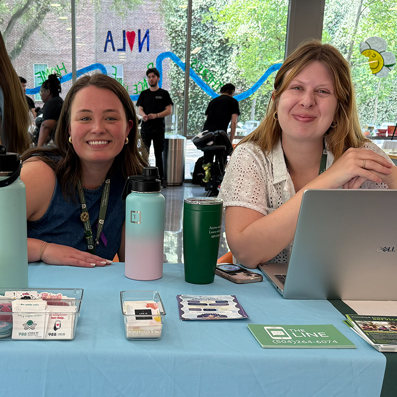 Two young women smile behind a table with water bottles and a laptop.