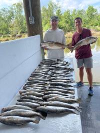 Two students smiling and holding large fish next to table full of caught fish
