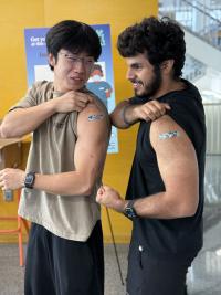 Two students smile and flex their arms next to each other, showing off bandages on their biceps from their flu shots
